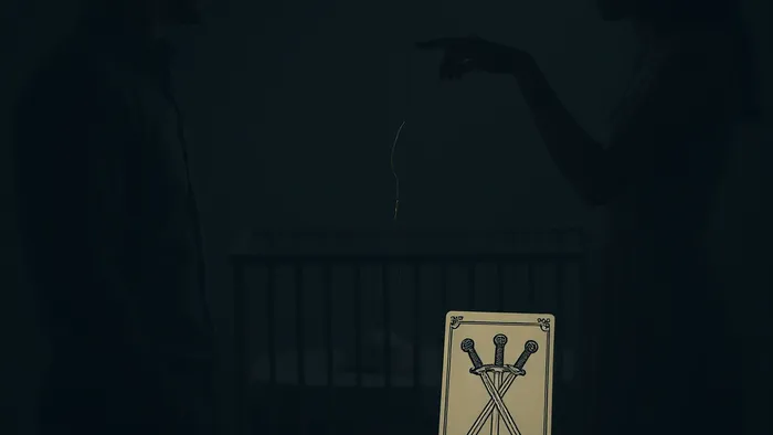 Символическое изображение пары в комнате с детской кроваткой между ними. На столе лежит карта Таро Двойка Мечей. Тонкая нить света тянется между ними, передавая ощущение хрупкости связи после рождения ребенка.